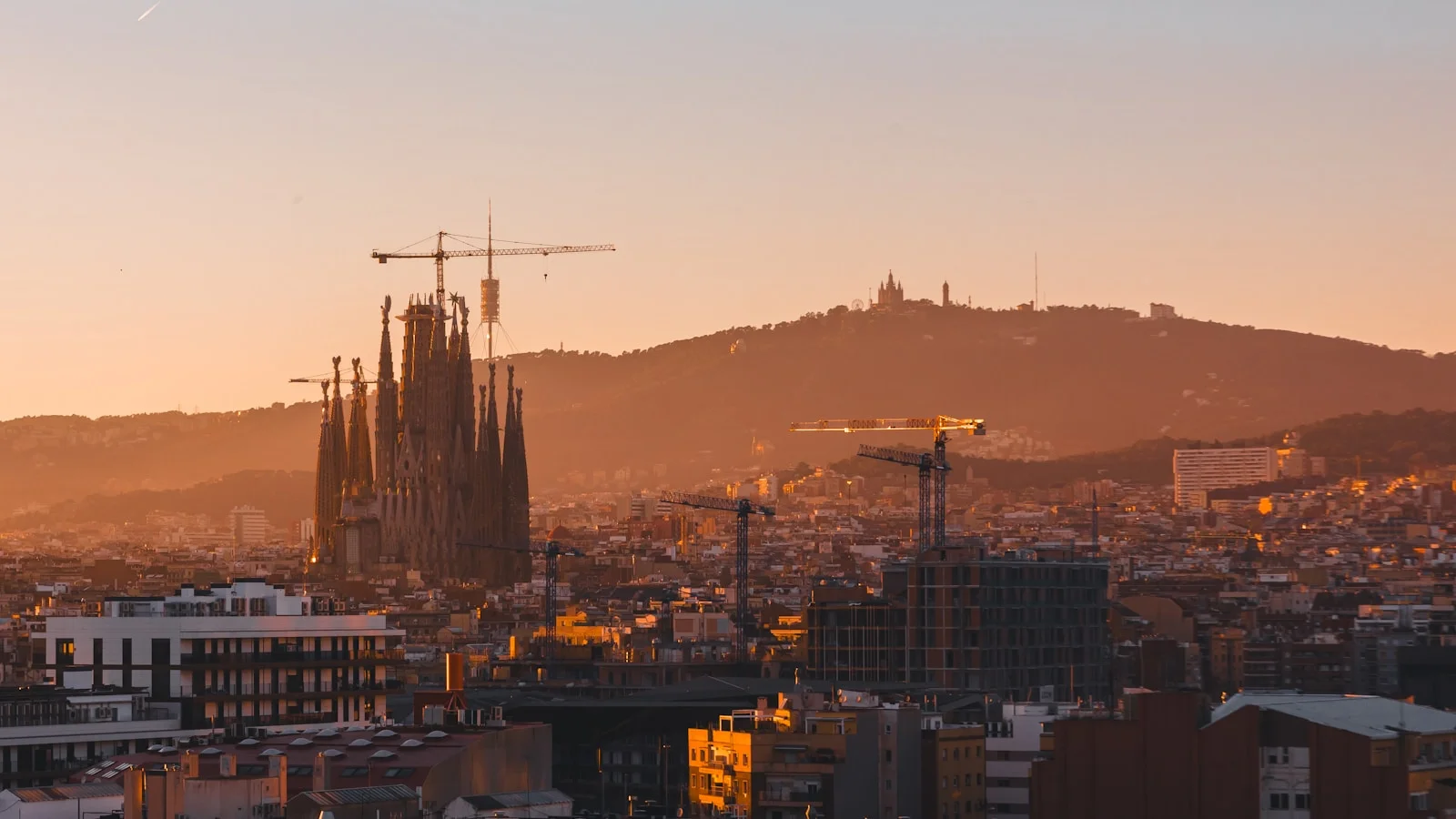 Barcelona skyline at sunset, with the unfinished spires of Sagrada Família rising above the dense Eixample grid and the Mediterranean behind.