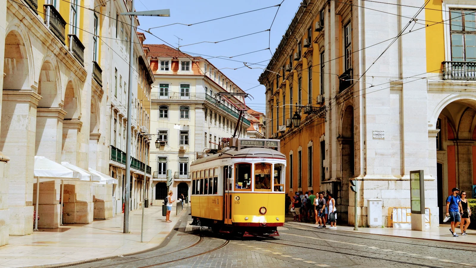 A yellow heritage tram climbing a steep cobblestone street in Lisbon's Alfama district, framed by pastel-tiled facades and laundry hanging from wrought-iron balconies.